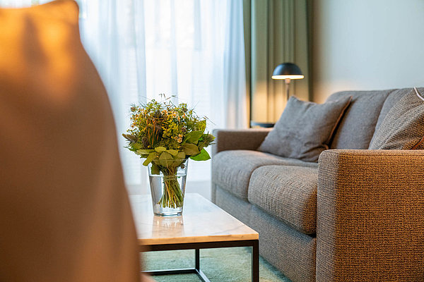 Der Wohnbereich eines FreiBlick Zimmers mit grünem Teppich, einer Couch und einer Blumenvase auf dem Tisch.
